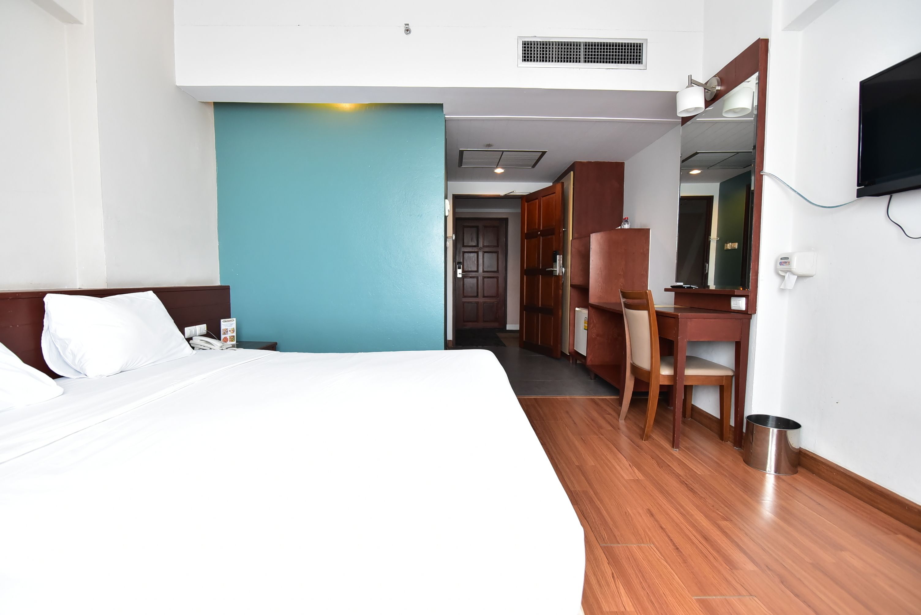 A hotel room with a large bed, a desk, and a chair. There is a TV mounted on the wall and a trash can on the floor.