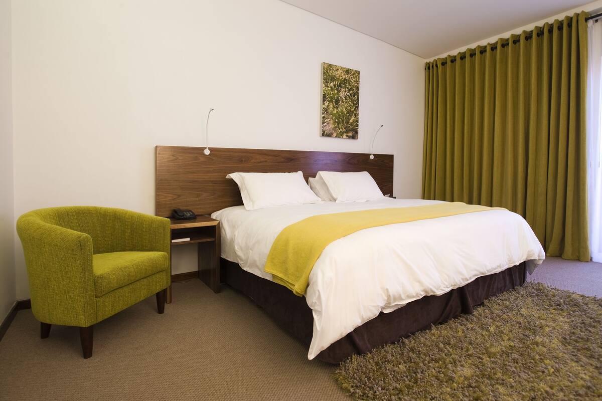Een hotelkamer met een bed, een groene fauteuil, een nachtkastje en een groot raam met groene gordijnen.