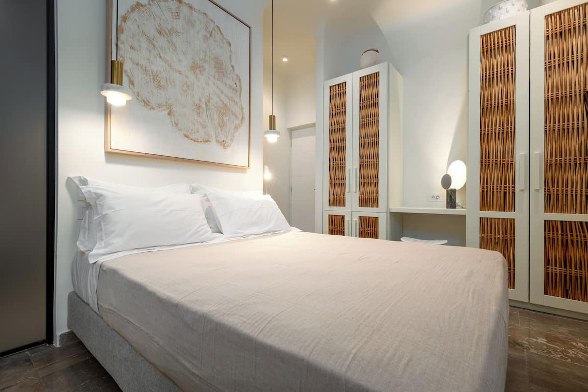 A neatly made bed in a modern room with a large abstract painting, a white wardrobe with a woven design, and a small table with a lamp.