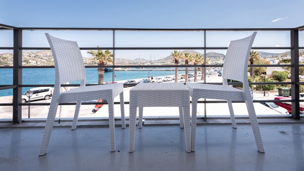 A balcony with two white wicker chairs and a table, overlooking a marina with boats and palm trees. - Hotel Oasis (Paros)