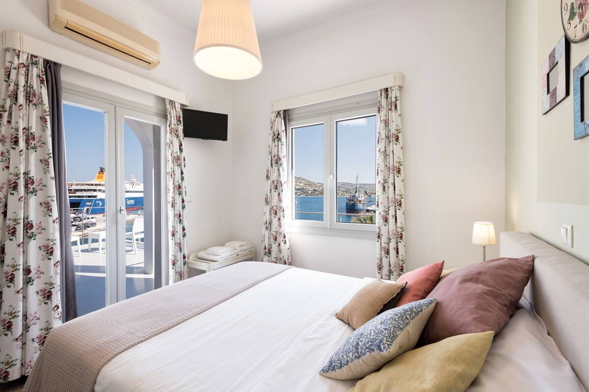 A hotel room with a bed, curtains, a TV, a window with a view of the sea, and a lamp.
