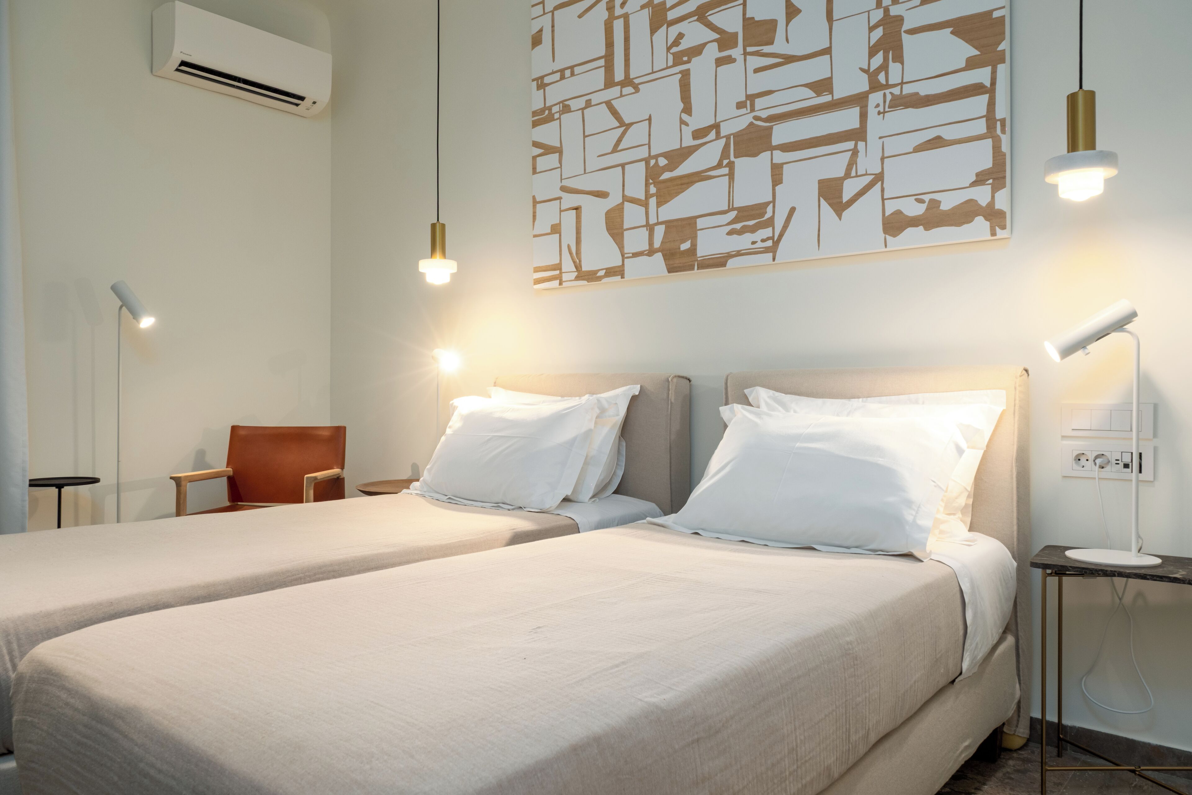 Habitación de hotel con dos camas, una silla marrón, aire acondicionado instalado en la pared y un cuadro abstracto moderno.