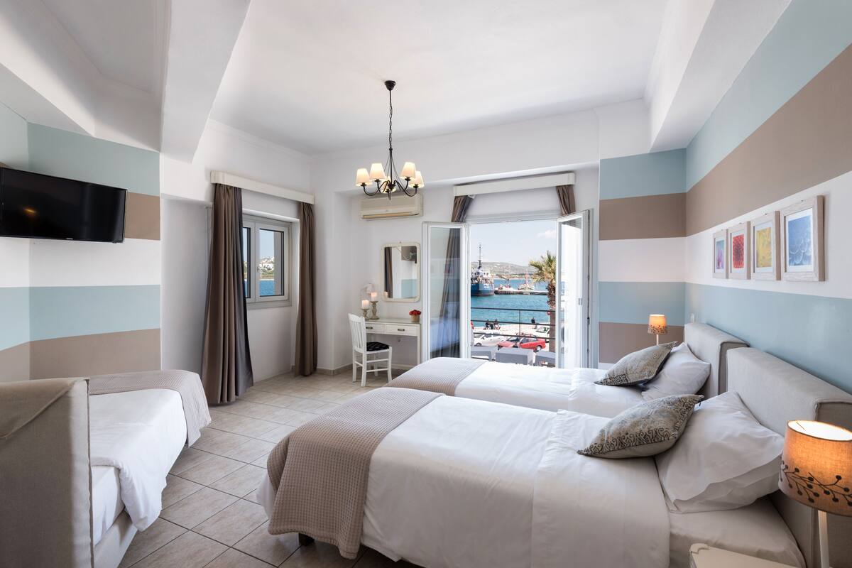 A hotel room with two beds, a desk, a TV, and a balcony with a view of the sea.