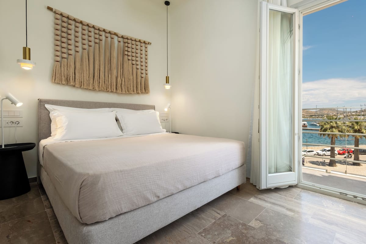 A bedroom with a bed, bedside lamps, a wall-mounted macrame piece, and a view of the sea through an open door.