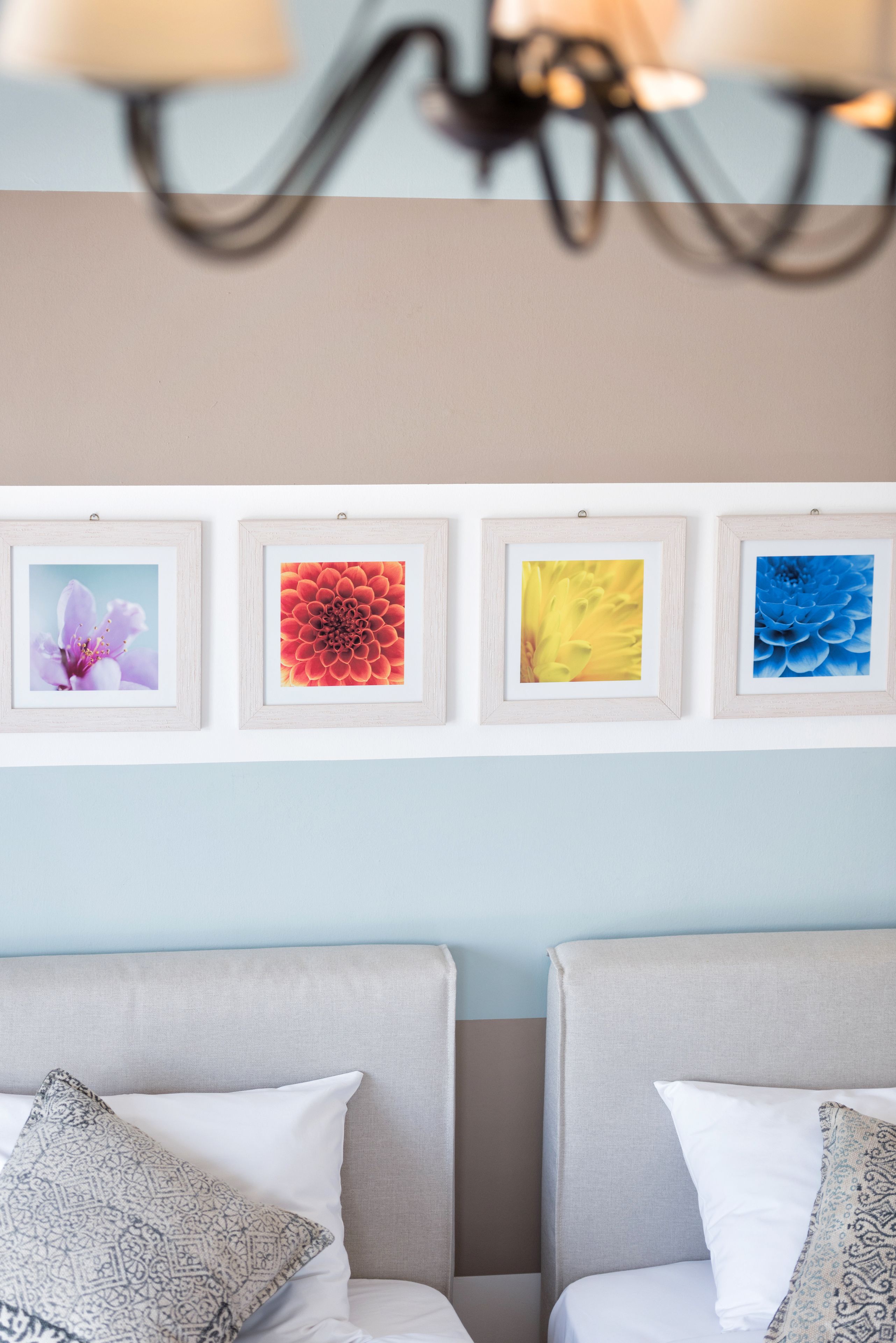 Una habitación con dos camas, una lámpara fijada a la pared y cuatro cuadros con motivos florales.