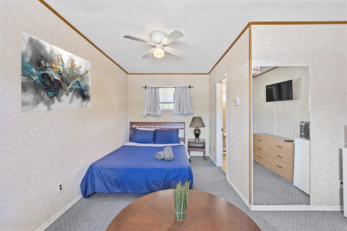 A bedroom with a bed, a ceiling fan, a painting, a window with curtains, a bedside table, and a mirror.