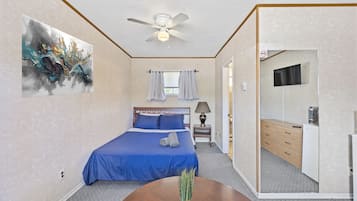 A bedroom with a bed, a ceiling fan, a painting, a window with curtains, a bedside table, and a mirror.