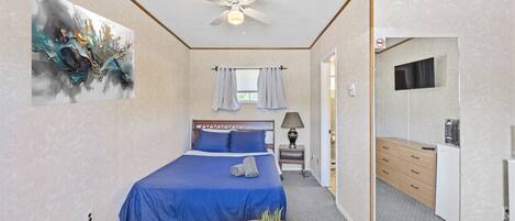 A bedroom with a bed, a ceiling fan, a painting, a window with curtains, a bedside table, and a mirror.