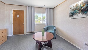 A room with a wooden door, a window with curtains, a blue armchair, a round table with a vase, and a wall-mounted abstract painting.