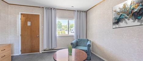 Una habitación con puerta de madera, ventana con cortinas, un sillón azul, una mesa redonda con un jarrón y un cuadro abstracto colgado en la pared.