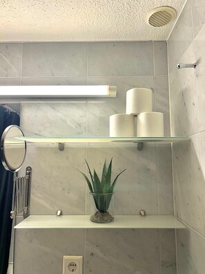 Een badkamer met marmeren tegels, een glazen plankje met een plant, een spiegel en toiletpapier.