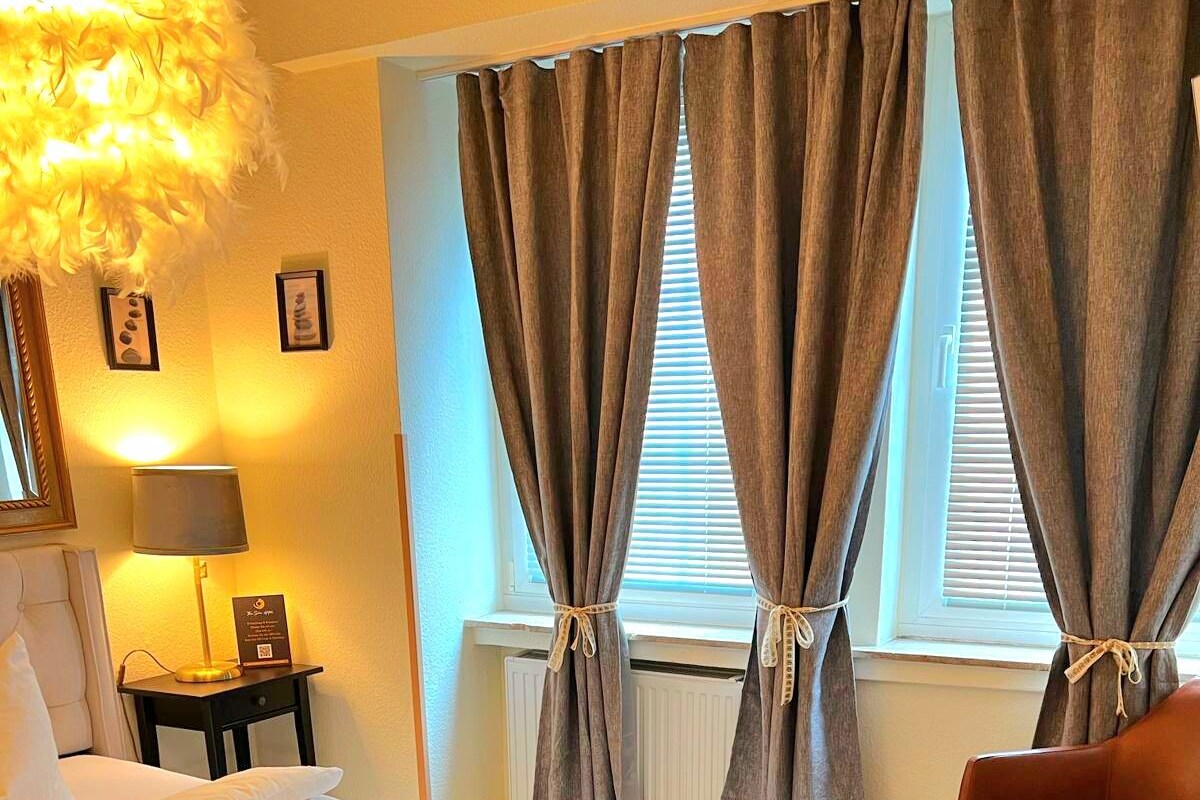 Een netjes opgemaakt bed met een donkere deken, een lamp op een nachtkastje en een stoel in een kamer met grote ramen en gordijnen.