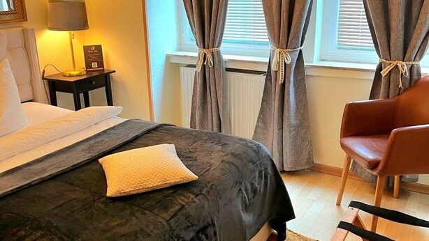 Een netjes opgemaakt bed met een donkere deken, een lamp op een nachtkastje en een stoel in een kamer met grote ramen en gordijnen.