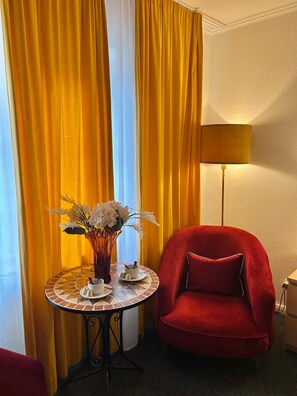 Ein Zimmer mit gelben Vorhängen, einem roten Sessel, einem Tisch mit Blumenvase und einer Lampe.