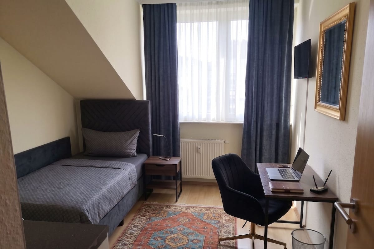 Een slaapkamer met een bed, een bureau met een laptop, een stoel, een radiator en een raam met gordijnen.