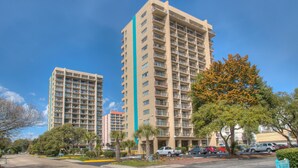 A high-rise hotel with multiple floors, balconies, and a parking area with cars. - The Dunes Towers by Palmetto Vacations (Myrtle Beach)
