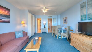 A living room with a sofa, coffee table, TV, and a dining area with chairs and a table. - The Dunes Towers by Palmetto Vacations (Myrtle Beach)
