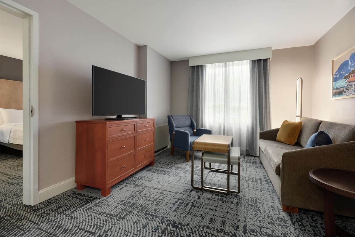 Habitación de hotel con televisor de pantalla plana, cómoda de madera, sillón azul, sofá gris con cojín mostaza y una pequeña mesa de madera.