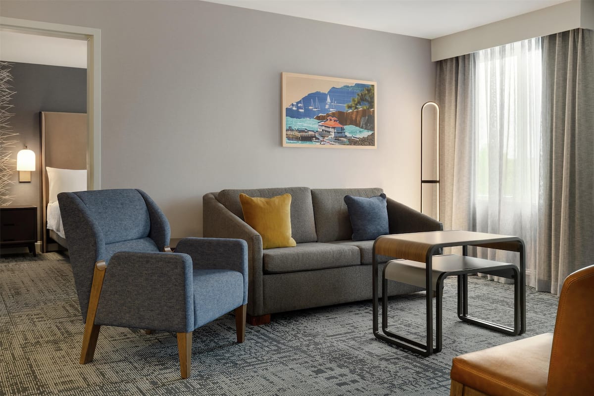 Una habitación de hotel moderna con un sofá gris, dos sillones, una mesita de madera y un cuadro de una escena costera.