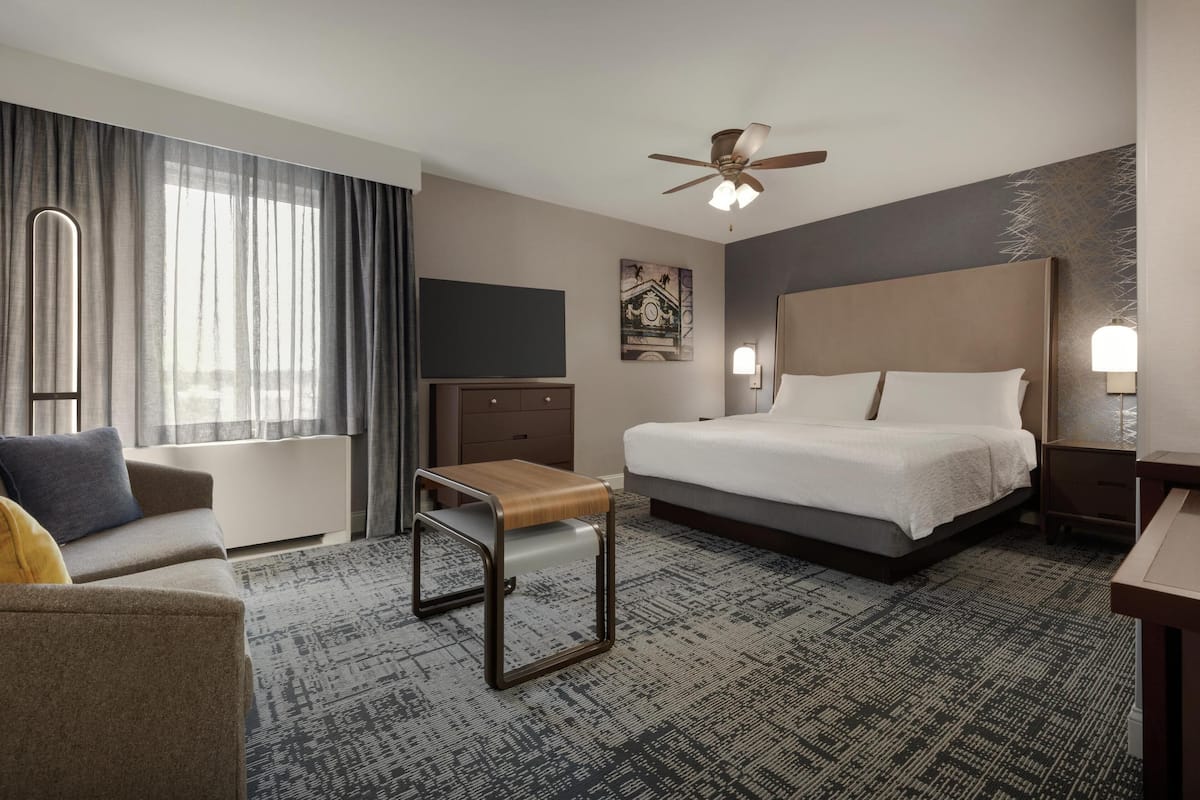 Habitación de hotel con cama, escritorio, silla, mesita pequeña, ventilador de techo y un amplio ventanal con cortinas.