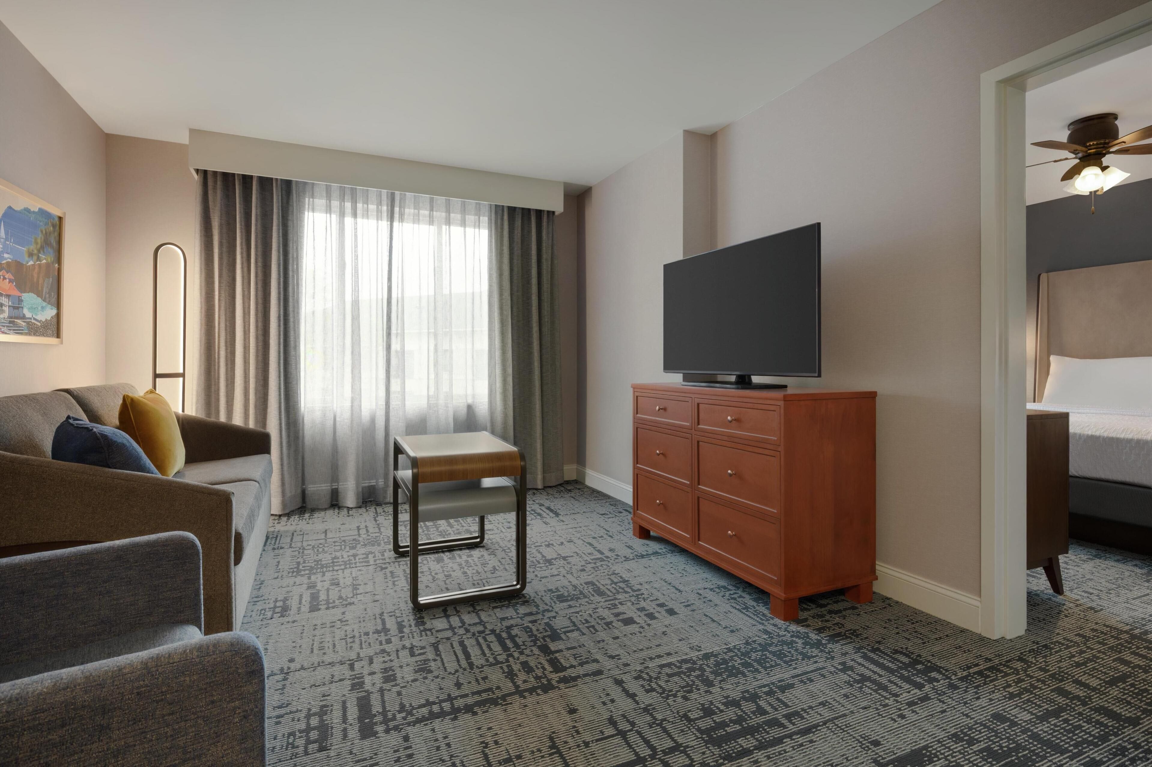 Ein Hotelzimmer mit einer Couch, einem kleinen Tisch, einem Fernseher auf einer Holzkommode und einem Bett mit Kopfteil.