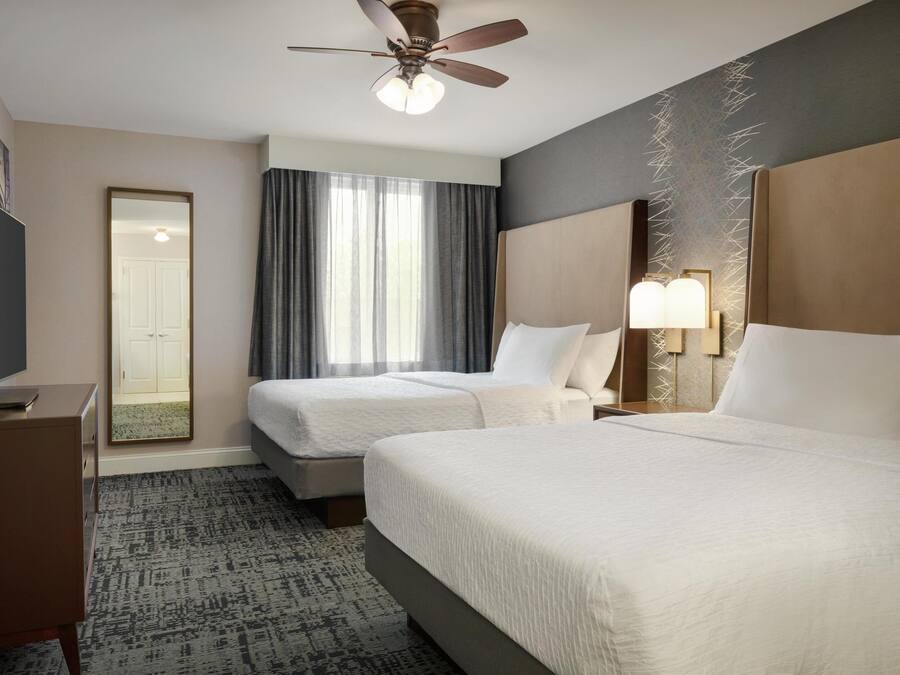 A hotel room with two beds, a ceiling fan, a TV, and a large window with curtains.