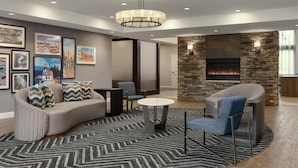A modern living room with a fireplace, comfortable seating, and framed cityscape pictures on the wall. - Homewood Suites by Hilton Burlington (South Burlington)