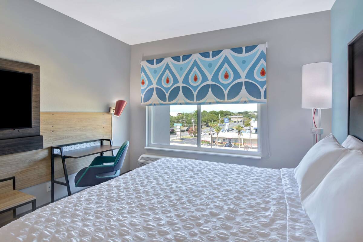 A hotel room with a large bed, a desk, a chair, a TV, a window with a view of the city, and a patterned curtain.