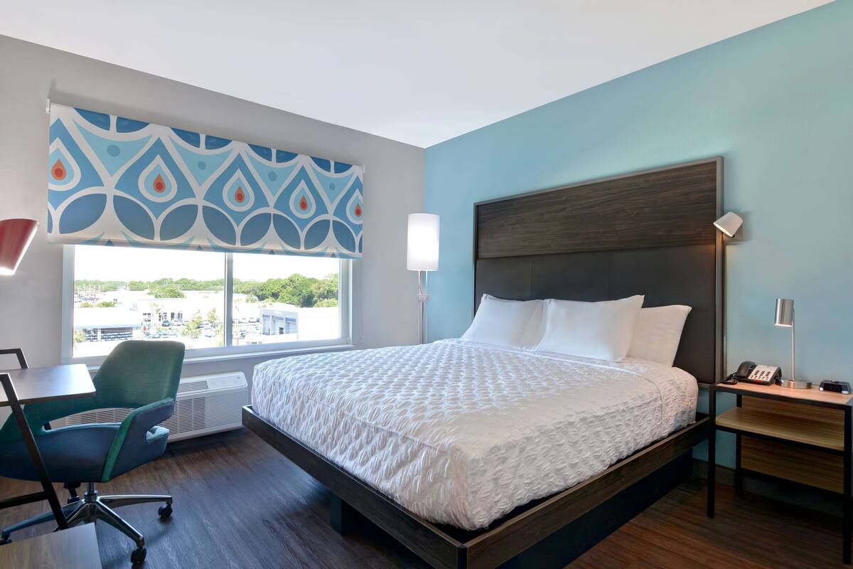 A hotel room with a large bed, a desk with a chair, a window with a view, and a bedside table with a lamp.