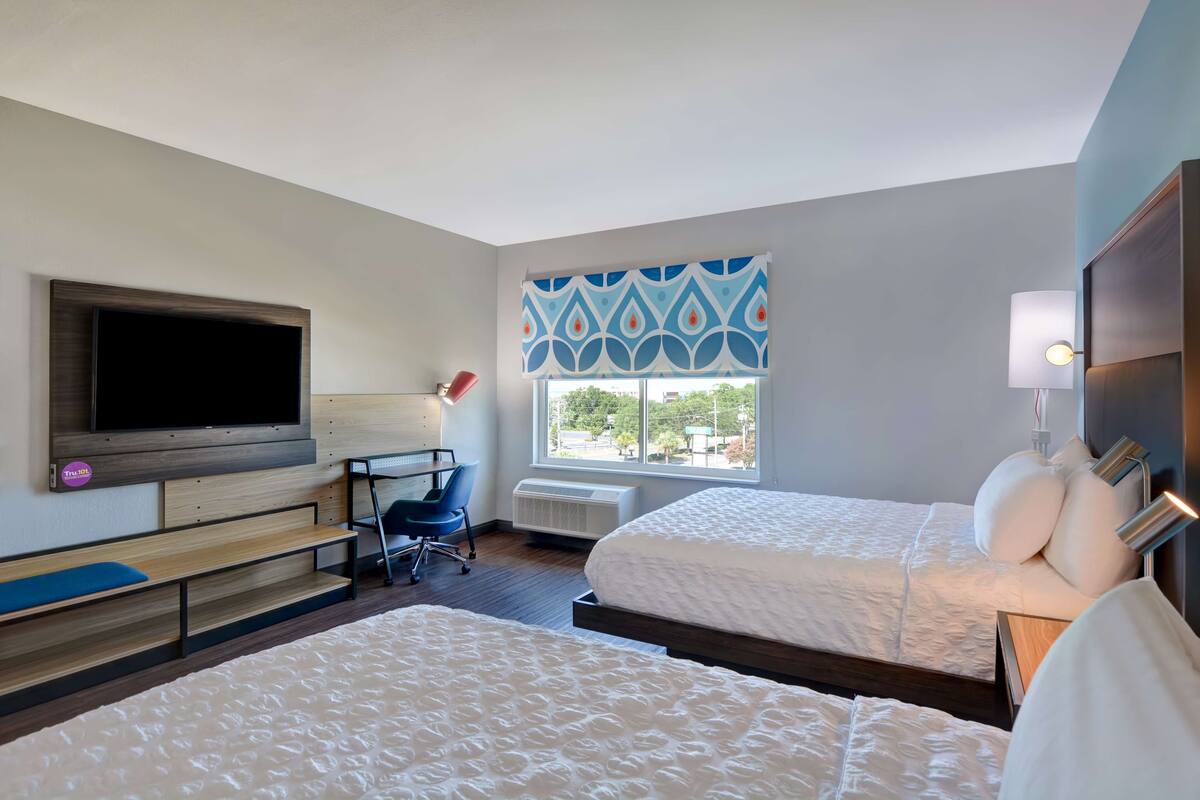 A hotel room with two beds, a TV mounted on the wall, a desk with a chair, and a window with patterned curtains.