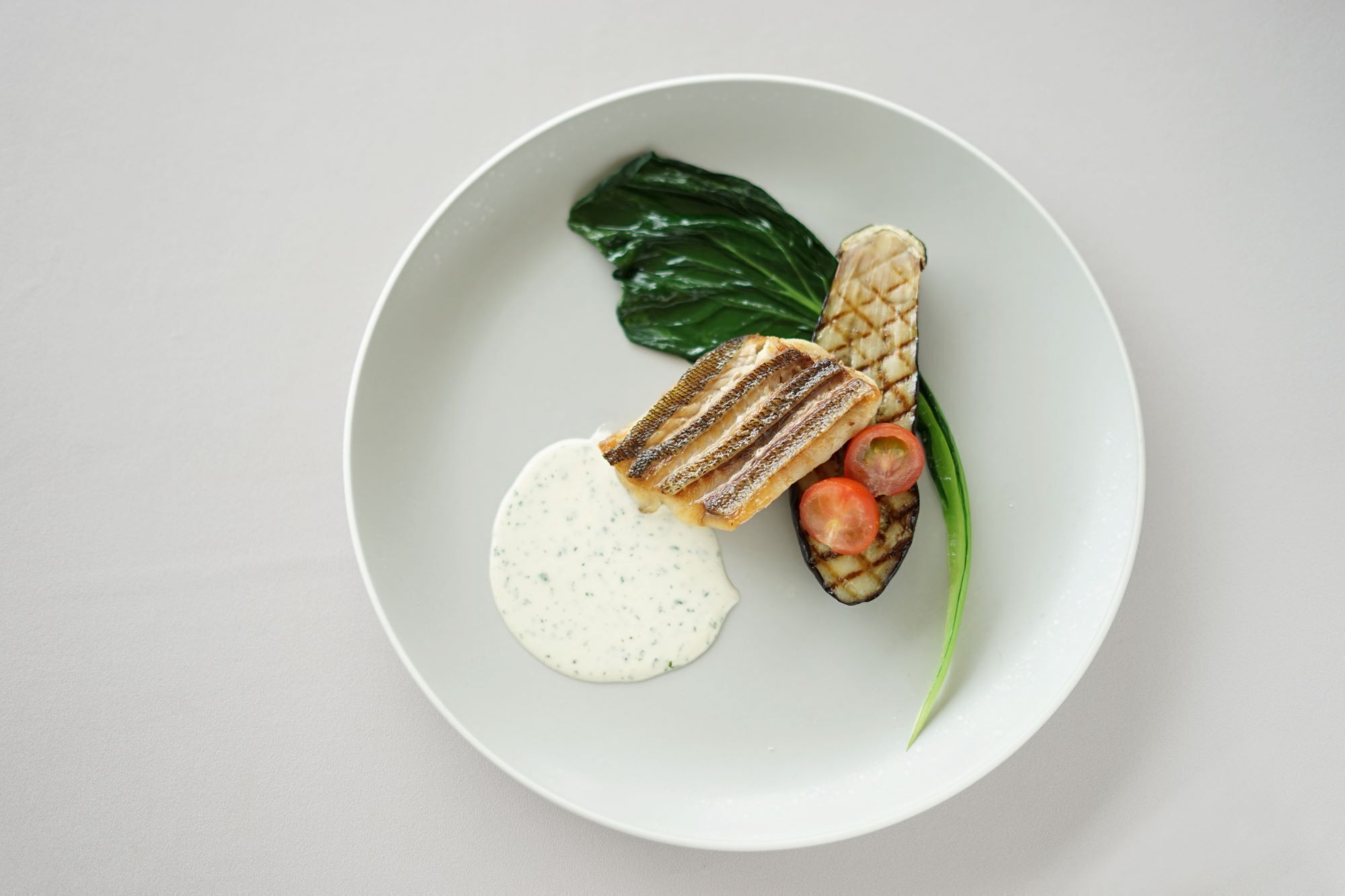 Un plato con pescado a la parrilla, una rodaja de tomate, una hoja de espinaca y una cucharada de salsa.