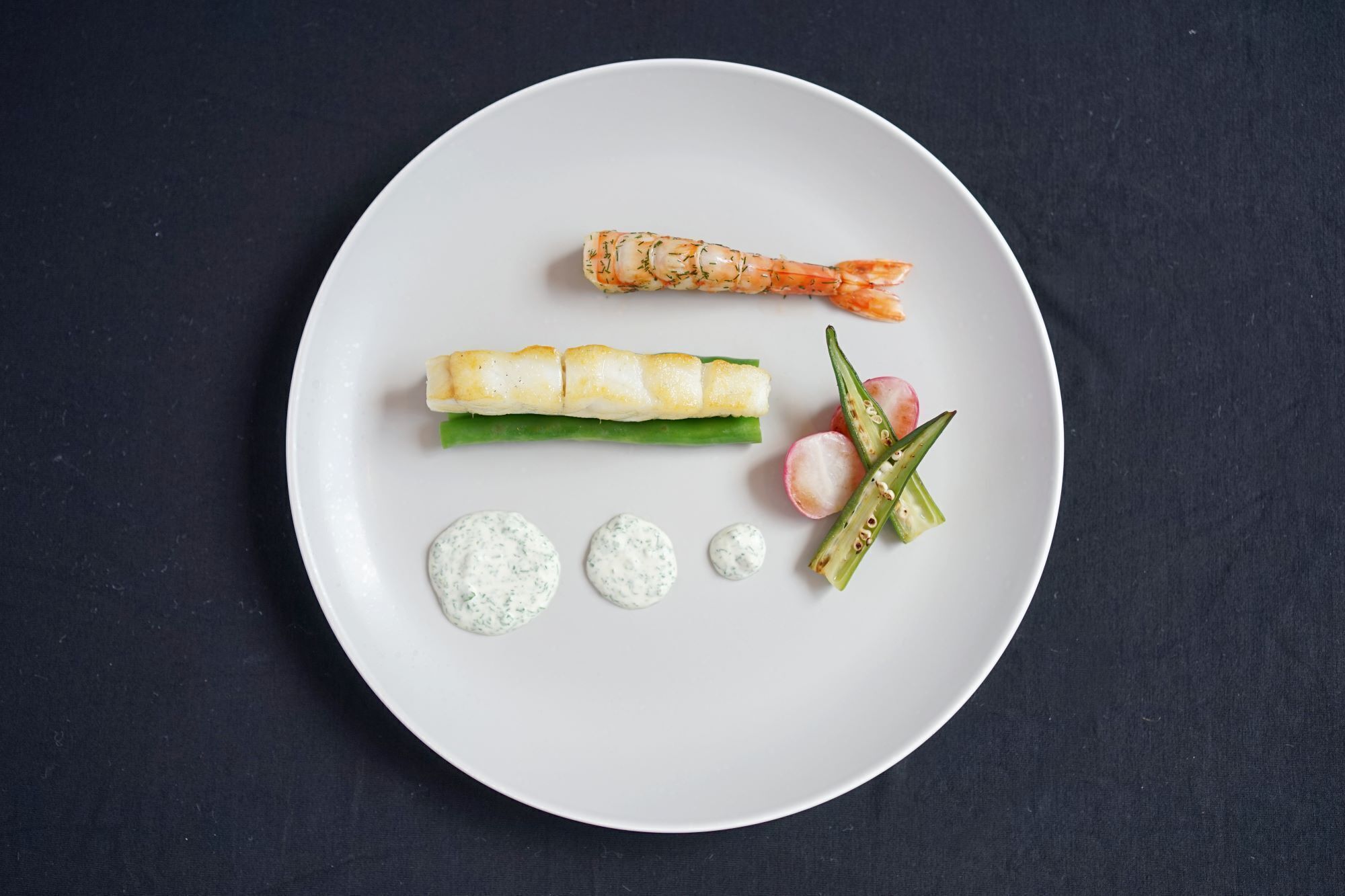 A white plate with a shrimp, a rolled fish, green beans, radishes, and two dollops of sauce.