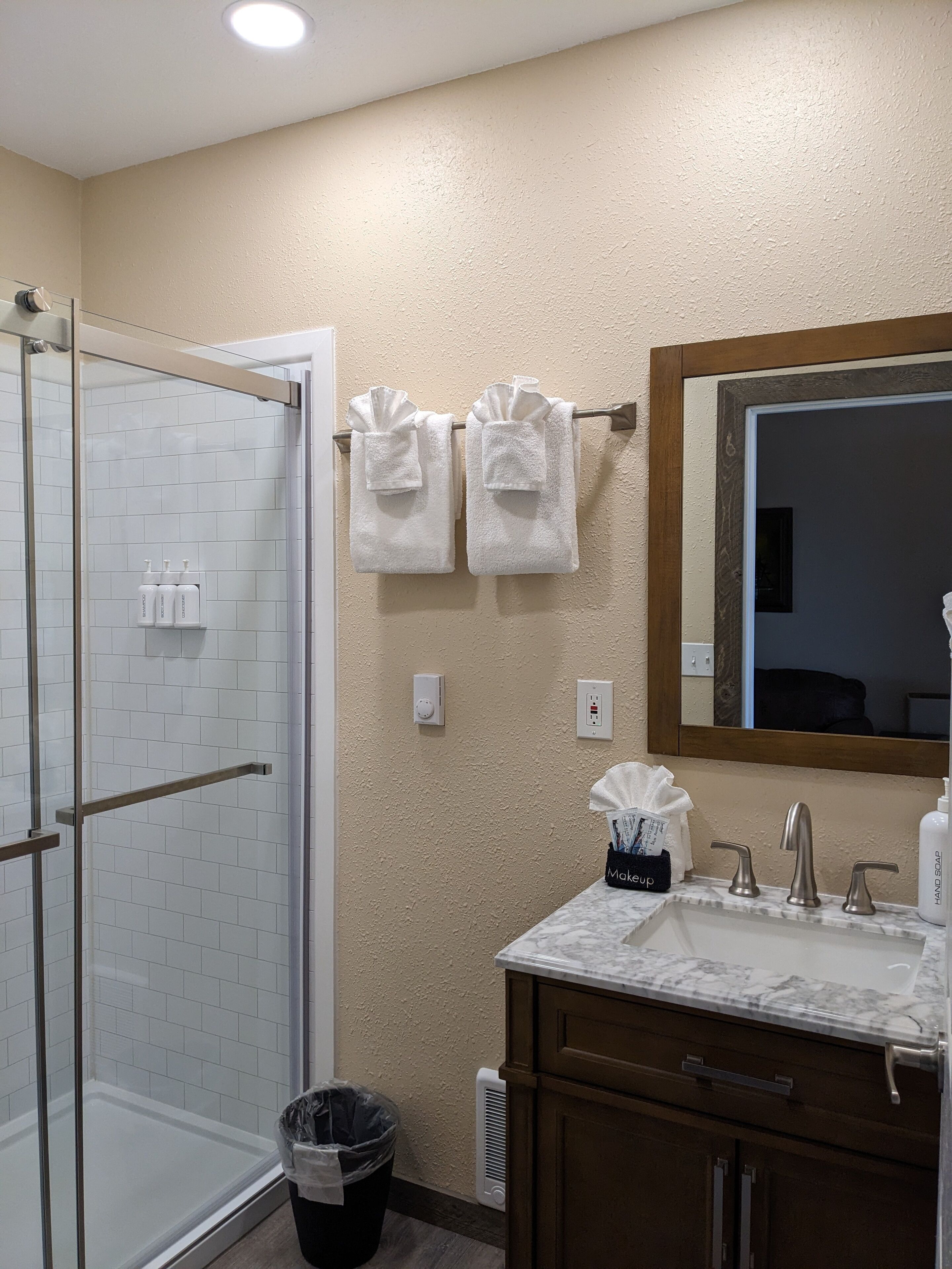 Un baño con mampara de vidrio, paredes de azulejos blancos, tocador de madera con encimera de mármol, un espejo y dos toallas blancas colgadas en un perchero.