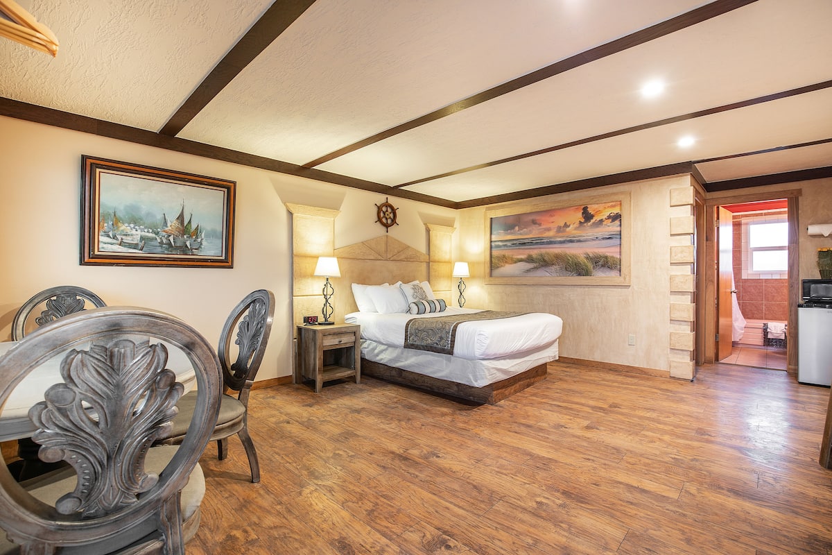 Un dormitorio con piso de madera, una cama con ropa blanca, una mesita de noche, dos lámparas de pared y dos cuadros.