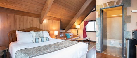 Un dormitorio amplio con una cama grande, vigas de madera en el techo, una zona de estar con sofá y sillones, y un baño con ducha e inodoro.