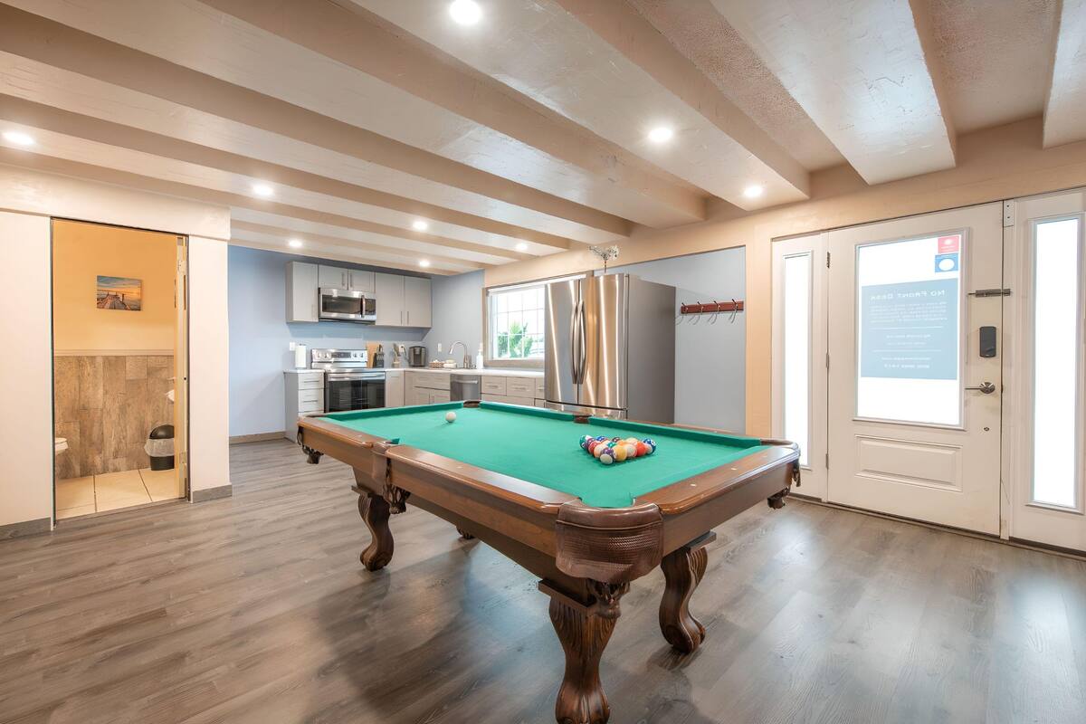 Una mesa de billar en una habitación con piso de madera, un área de cocina y una puerta con un letrero.