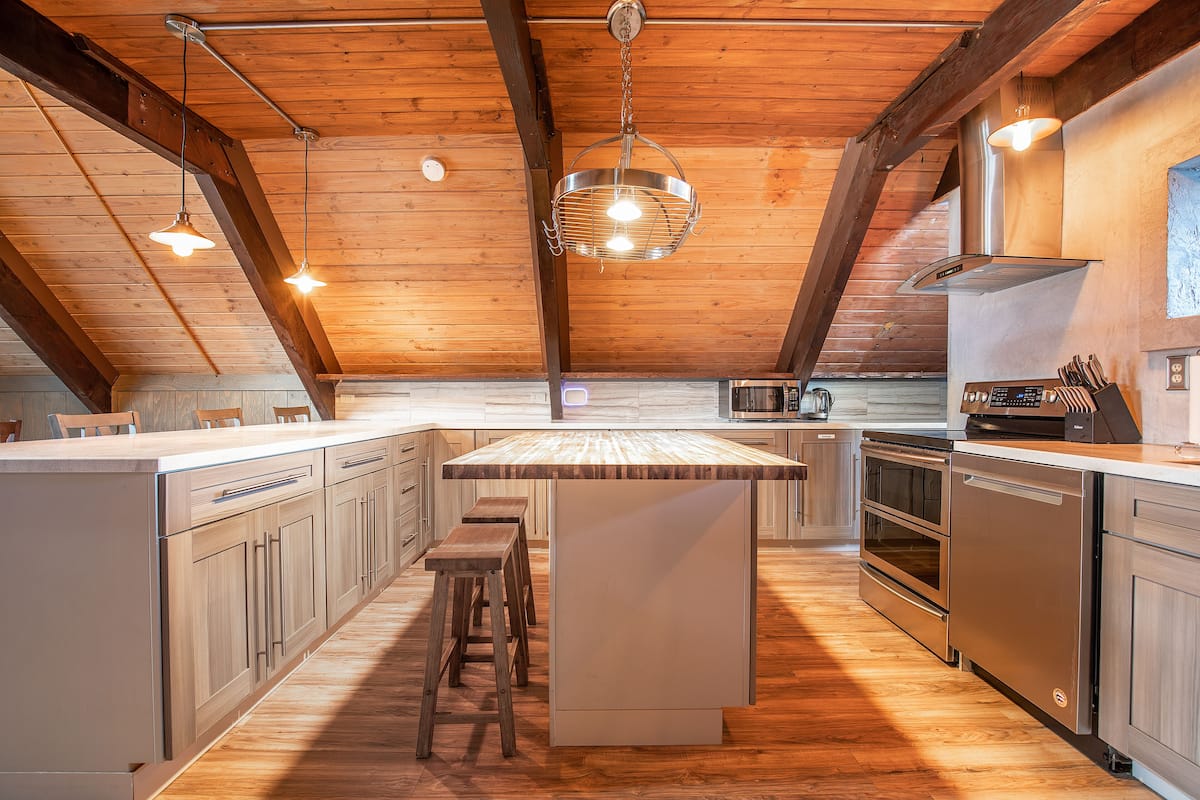 Una cocina con vigas de madera, una isla grande con taburetes, electrodomésticos de acero inoxidable e iluminación colgante.