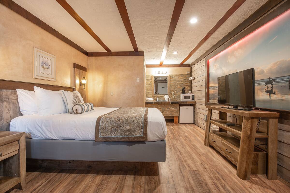 Un dormitorio con cama, cabecero de madera, televisor sobre un soporte de madera, baño con ducha y un amplio ventanal con vista a la playa.