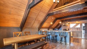 A high-ceilinged dining area with wooden tables and benches, a kitchen with stainless steel appliances, and a view of the outdoors through a window. - Wanderlust Inn (Ocean Shores)