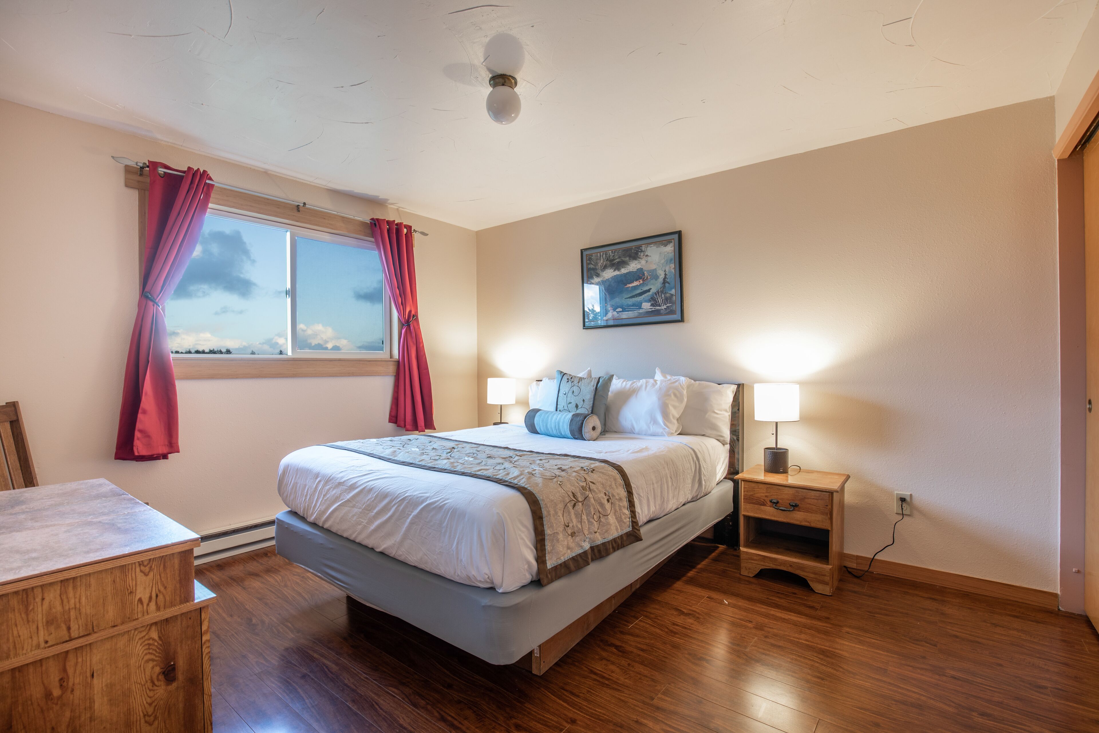 Un dormitorio con cama, mesitas de noche, una ventana con cortinas, un cuadro en la pared y suelo de madera.