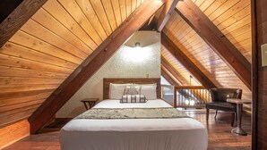 A cozy attic bedroom with a wooden sloped ceiling, a large bed with a decorative headboard, a small table, and a chair. - Wanderlust Inn (Ocean Shores)