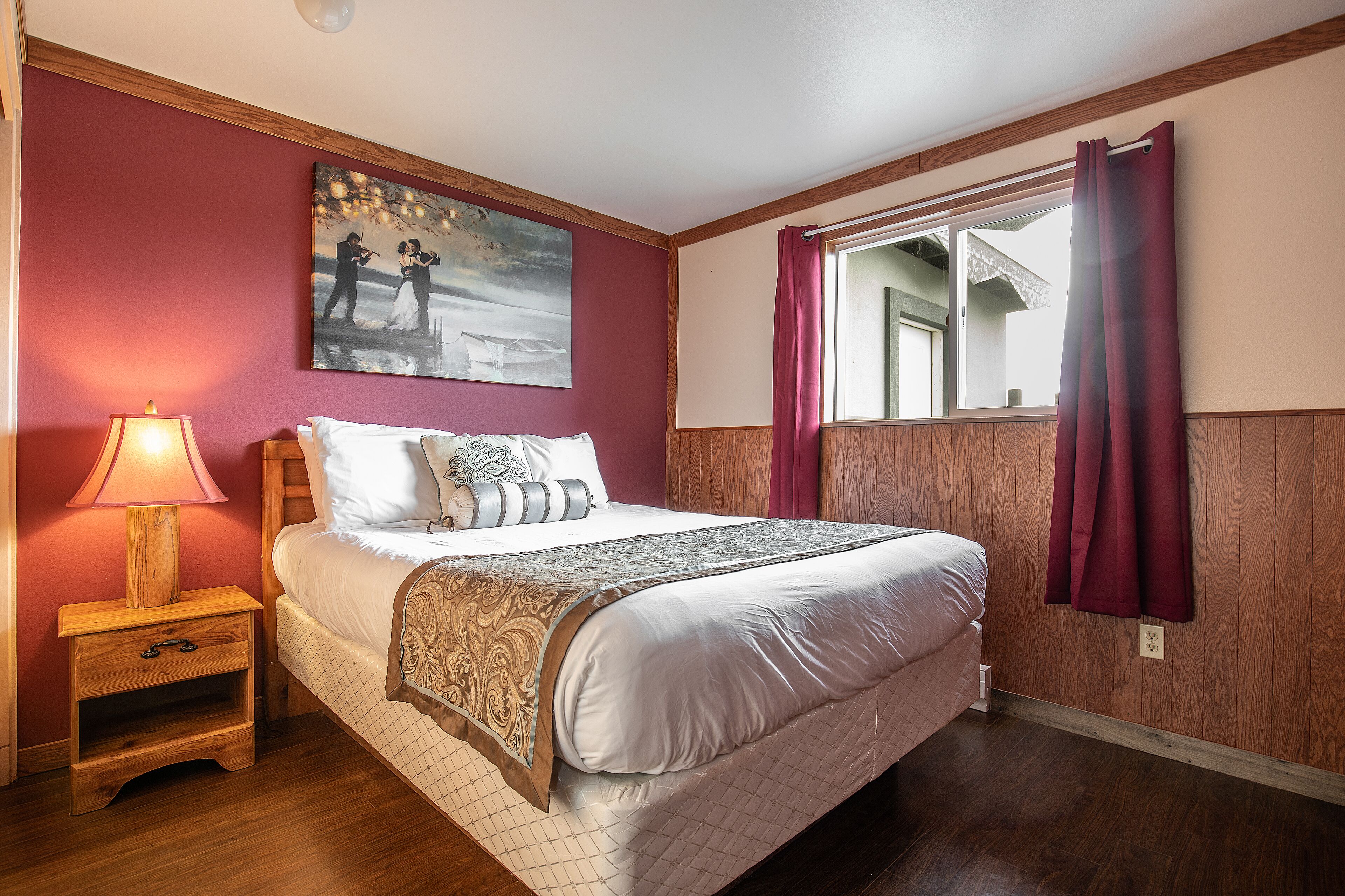 A bedroom with a bed, a nightstand, a lamp, a painting, a window with curtains, and a view of the outside.