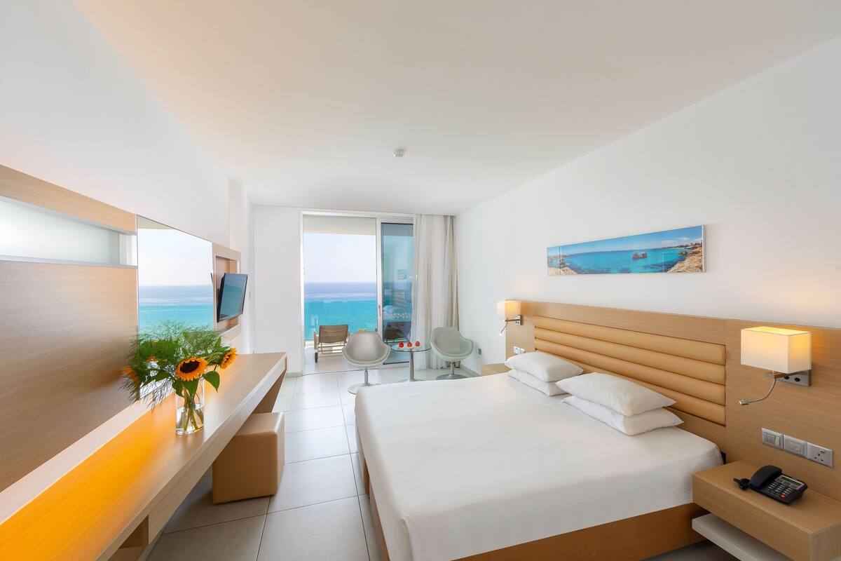 A modern hotel room with a large bed, a desk with a vase of flowers, a chair, a small table, and a view of the ocean.