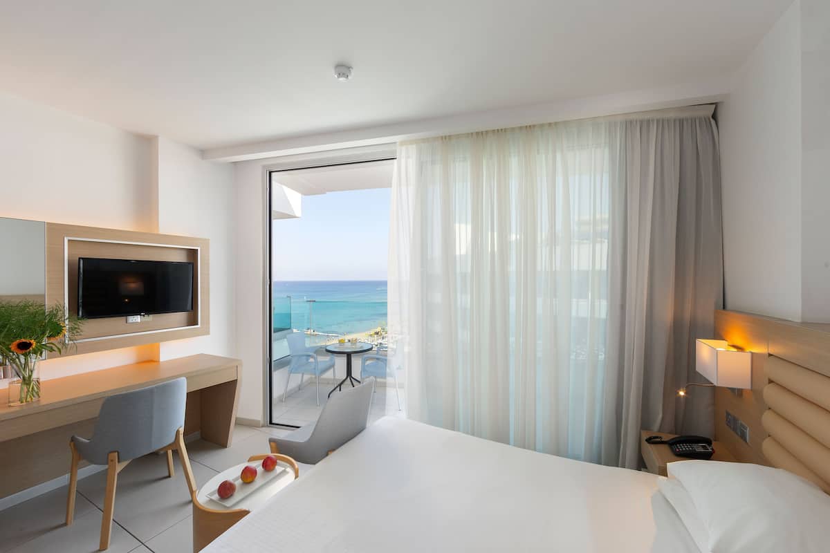 A hotel room with a bed, a desk with a TV, a chair, a small table with fruit, a nightstand with a lamp, and a view of the ocean.