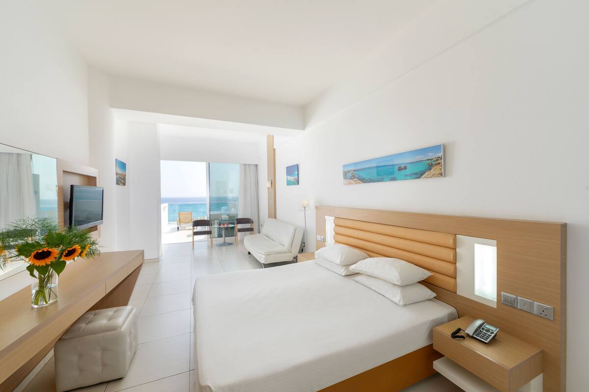 A modern hotel room with a large bed, a desk with a phone, a TV, and a view of the ocean.