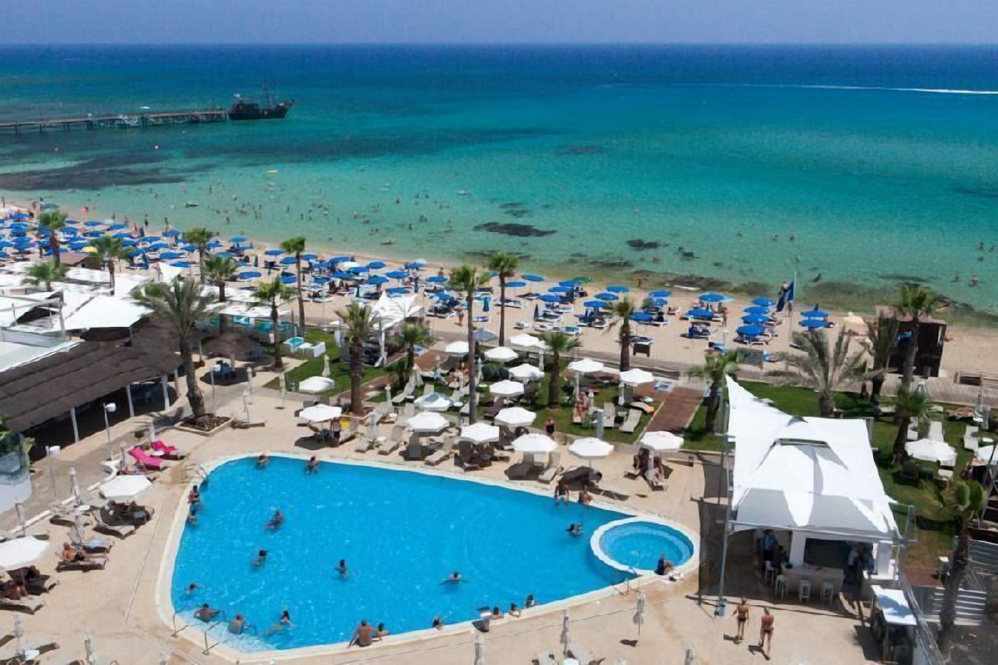 Amplia zona de piscina con numerosas tumbonas y sombrillas, rodeada de asientos frente a la playa y vistas al océano.