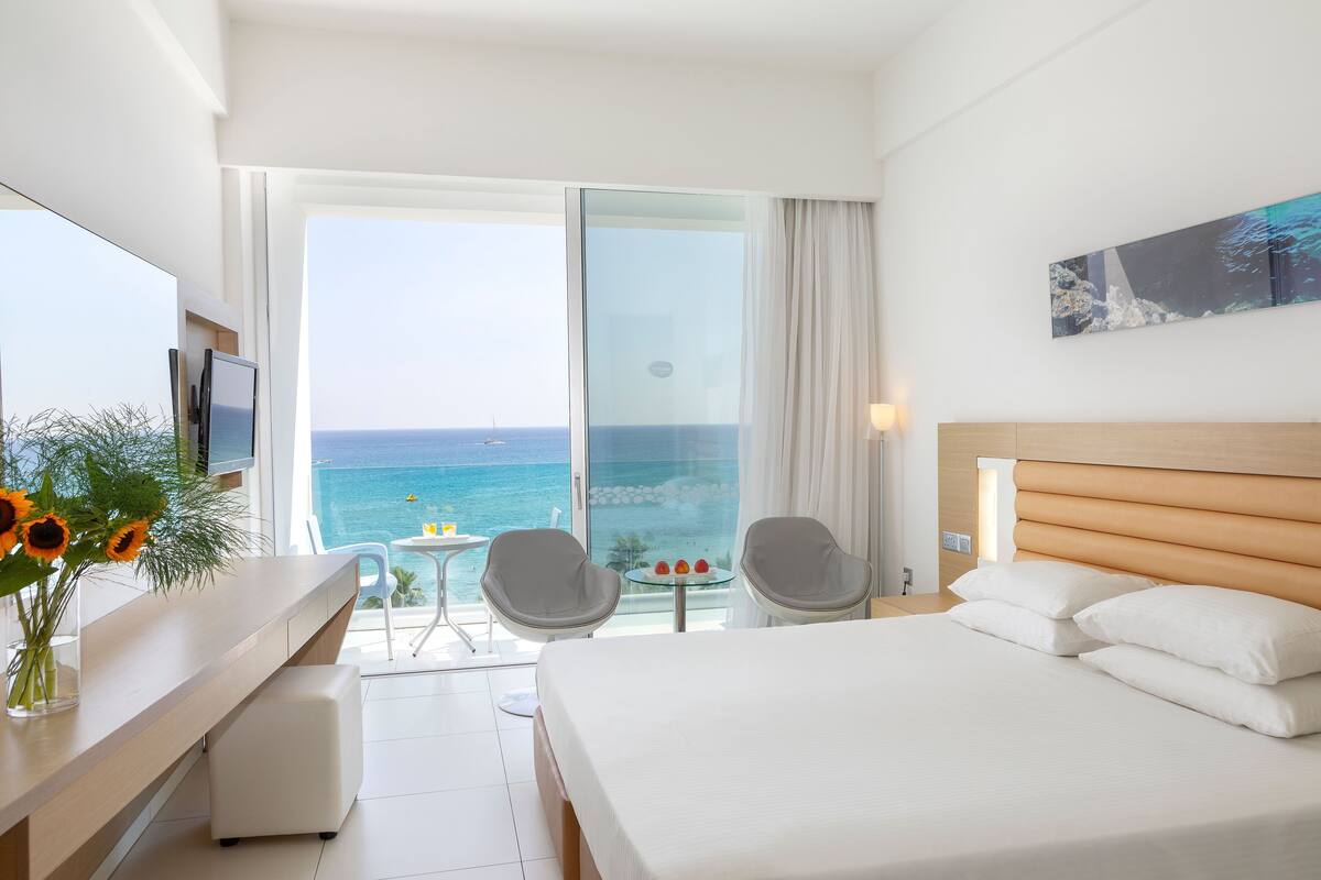 A hotel room with a bed, a desk, a chair, a small table with a vase of flowers, and a large window with a view of the ocean.
