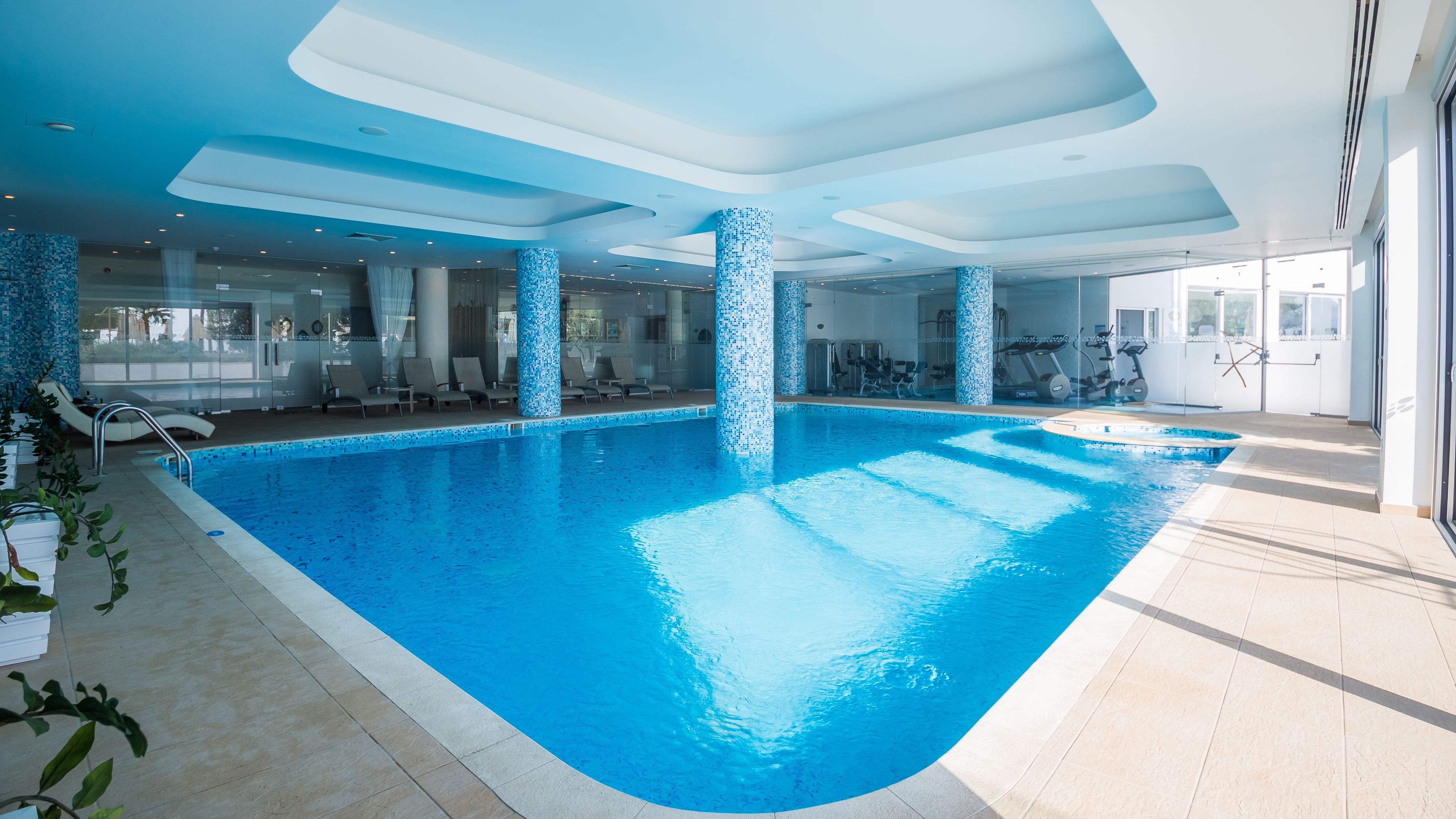 Ein Hallenbad mit klarem, blauen Wasser, umgeben von Liegestühlen und Fitnessgeräten.