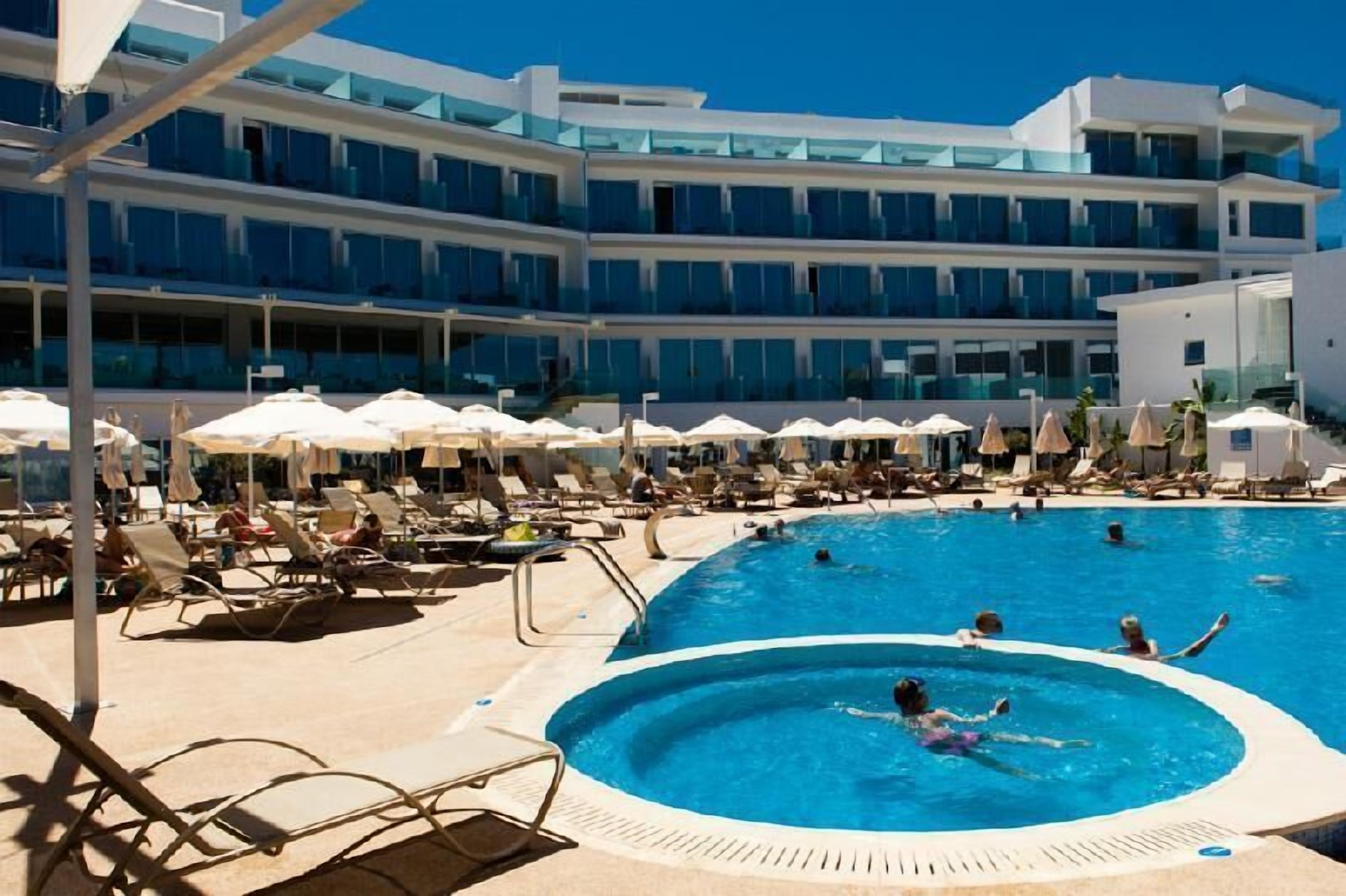 A pool area with lounge chairs and umbrellas, surrounded by a modern hotel building with multiple floors and balconies.