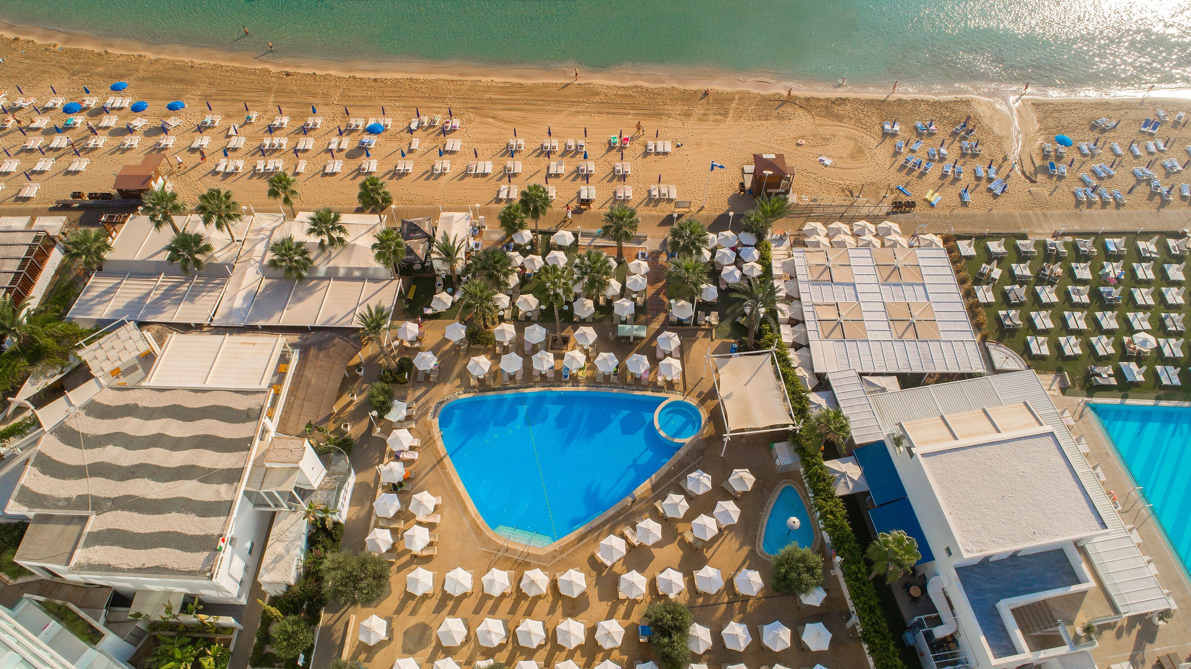 Vista aerea di un resort con una grande piscina, diverse zone sulla spiaggia con lettini e vari ombrelloni.